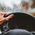 Close-up of a hand holding a steering wheel, with heavy rain hitting the windshield.