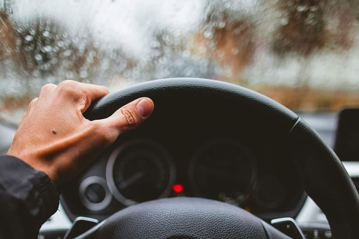 Close-up of a hand holding a steering wheel, with heavy rain hitting the windshield.